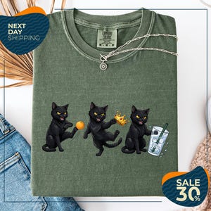 a green t-shirt with a graphic design of three black cats, each holding a different object: a ball, a crown, and a glass of water. The cats are depicted in a playful and whimsical manner, with one cat holding a crown and the other two holding a ball and a glass of water. The t-shirt is displayed on a white background, and there are some jeans and a pair of sunglasses visible in the background.