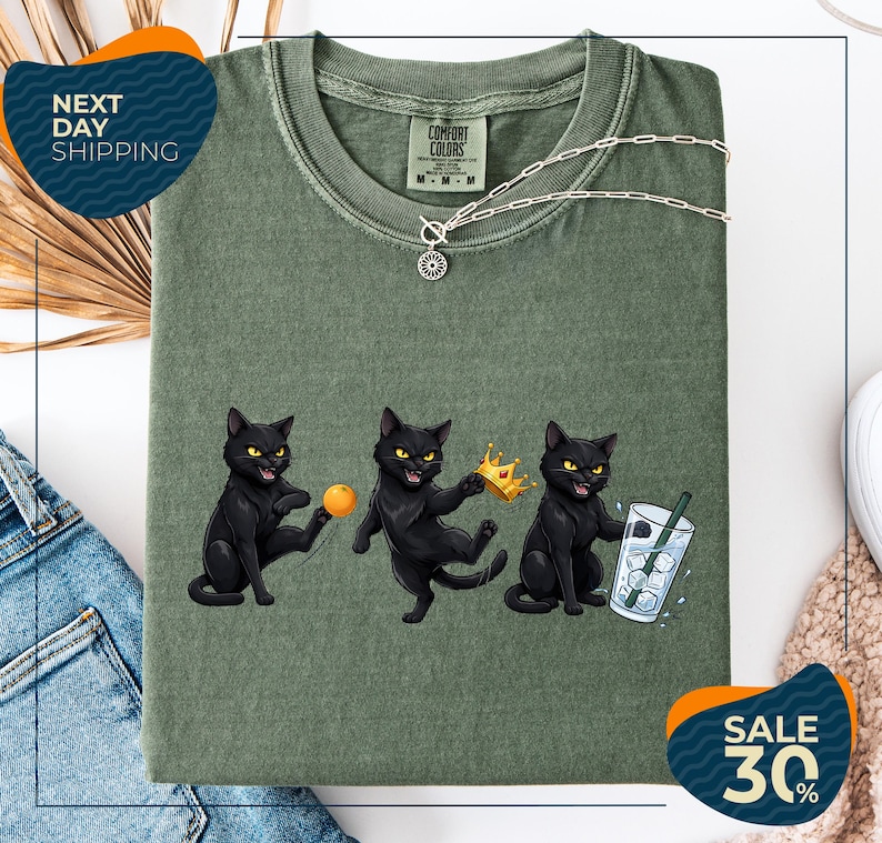 a green t-shirt with a graphic design of three black cats, each holding a different object: a ball, a crown, and a glass of water. The cats are depicted in a playful and whimsical manner, with one cat holding a crown and the other two holding a ball and a glass of water. The t-shirt is displayed on a white background, and there are some jeans and a pair of sunglasses visible in the background.