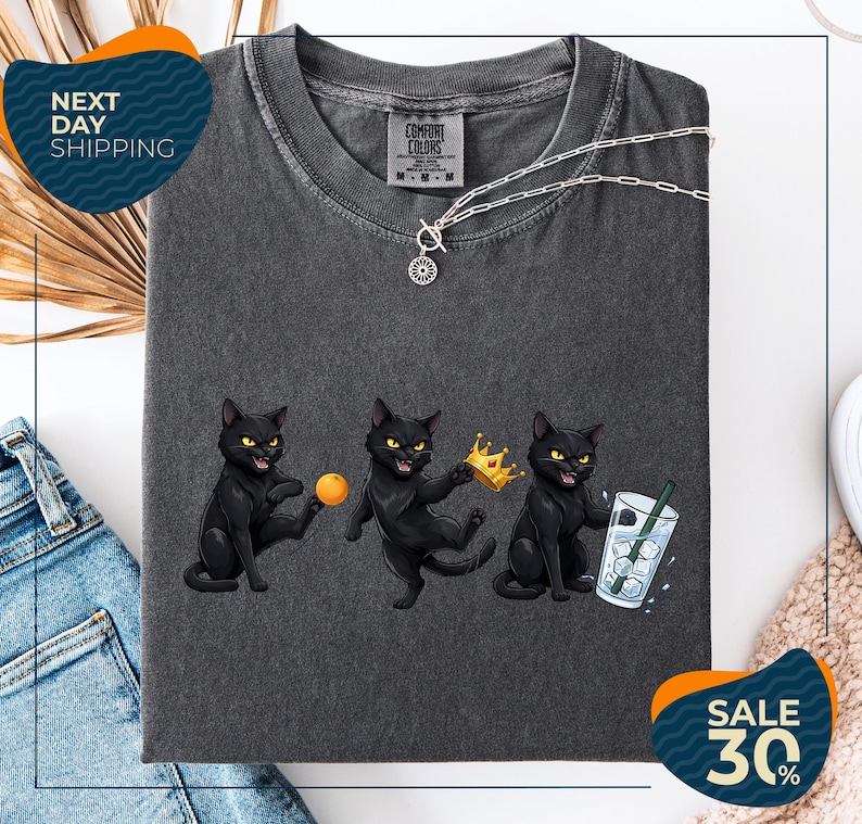 a gray t-shirt with a graphic design of three black cats, each holding a different object: a ball, a crown, and a drink. The t-shirt is displayed on a white background, and there are jeans and a white object in the background. The image also includes text that reads "Next Day Shipping" and "SALE 30%".