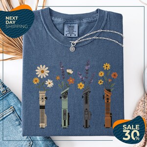 a blue t-shirt with a graphic design of four different colored vases holding flowers, arranged in a row. The vases are depicted in various colors, including blue, green, orange, and brown. The design also includes a small charm hanging from the t-shirt. The t-shirt is displayed on a white background, and there are some jeans and a beige sweater visible in the background.