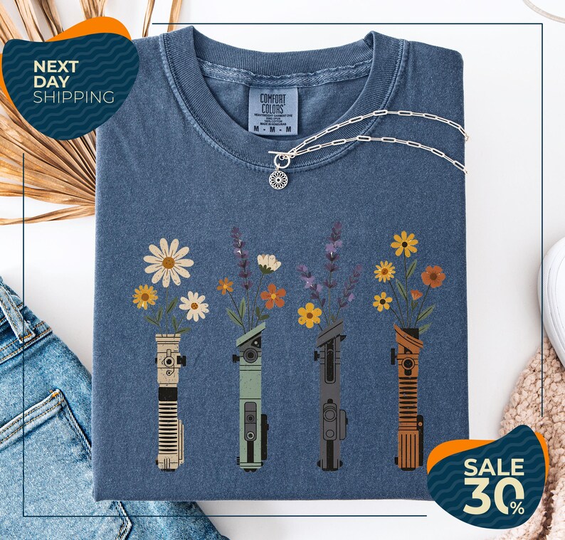 a blue t-shirt with a graphic design of four different colored vases holding flowers, arranged in a row. The vases are depicted in various colors, including blue, green, orange, and brown. The design also includes a small charm hanging from the t-shirt. The t-shirt is displayed on a white background, and there are some jeans and a beige sweater visible in the background.