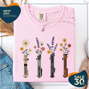 a pink t-shirt with a graphic design of four colorful vases holding flowers, arranged in a row. The vases are depicted in various colors, including pink, green, blue, and brown. The t-shirt is displayed on a white background, and there are some jeans and a necklace visible in the background.