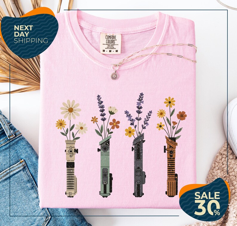 a pink t-shirt with a graphic design of four colorful vases holding flowers, arranged in a row. The vases are depicted in various colors, including pink, green, blue, and brown. The t-shirt is displayed on a white background, and there are some jeans and a necklace visible in the background.