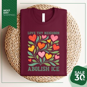 a burgundy t-shirt with a colorful graphic design of hearts and leaves, along with the text "Love thy neighbor" and "Abolish ice" printed on it. The t-shirt is displayed on a woven basket, and there is a green banner with white text that reads "Next day shipping" and "SALE 30%".