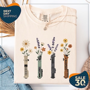 a white t-shirt with a graphic design of four colorful vases holding flowers, arranged in a row. The vases are of different colors and shapes, creating a visually appealing pattern. The t-shirt is displayed on a white background, and there are some jeans and a sweater visible in the background. The image also includes text that reads "Next Day Shipping" and "SALE 30%", indicating that the t-shirt is available for immediate shipping and is on sale at a discounted price.