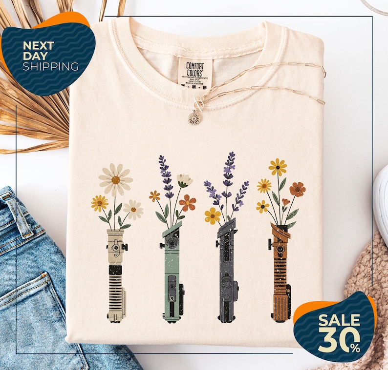 a white t-shirt with a graphic design of four colorful vases holding flowers, arranged in a row. The vases are of different colors and shapes, creating a visually appealing pattern. The t-shirt is displayed on a white background, and there are some jeans and a sweater visible in the background. The image also includes text that reads "Next Day Shipping" and "SALE 30%", indicating that the t-shirt is available for immediate shipping and is on sale at a discounted price.