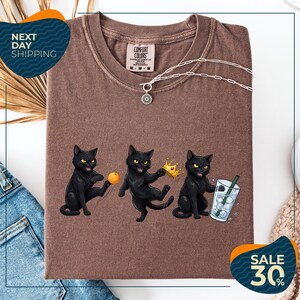 a brown t-shirt with a graphic design of three black cats, each holding a different object: a crown, a ball, and a glass of water. The cats are depicted in a playful and whimsical manner, with one cat holding a crown and the other two holding a ball and a glass of water. The t-shirt is displayed on a white background, and there are some jeans and a sweater visible in the background.