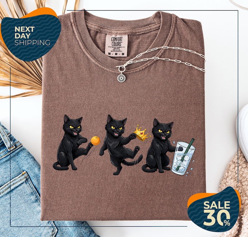 a brown t-shirt with a graphic design of three black cats, each holding a different object: a crown, a ball, and a glass of water. The cats are depicted in a playful and whimsical manner, with one cat holding a crown and the other two holding a ball and a glass of water. The t-shirt is displayed on a white background, and there are some jeans and a sweater visible in the background.