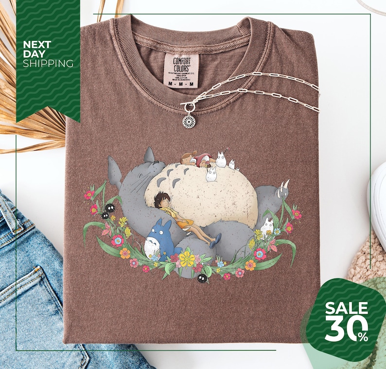 a brown t-shirt with a colorful graphic design depicting a cat surrounded by various animals and flowers. The t-shirt is displayed on a white background, and there is a sale sign indicating a 30% discount.
