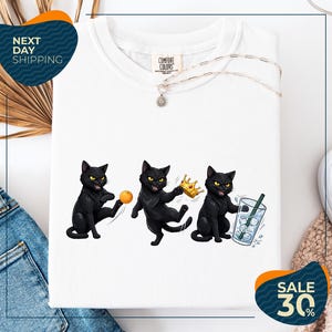 a white t-shirt with a graphic design of three black cats, each holding a different object: a ball, a crown, and a glass of water. The cats are depicted in various poses, creating a dynamic and playful scene. The t-shirt is displayed on a white background, and there are some jeans and a sweater visible in the background. The image also includes text that reads "Next Day Shipping" and "SALE 30%".
