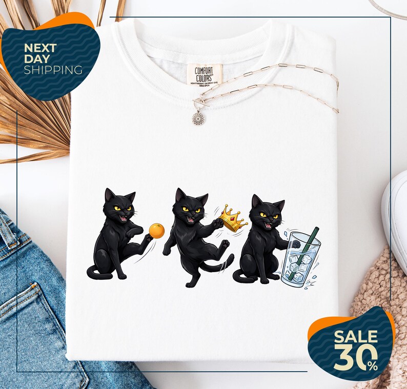 a white t-shirt with a graphic design of three black cats, each holding a different object: a ball, a crown, and a glass of water. The cats are depicted in various poses, creating a dynamic and playful scene. The t-shirt is displayed on a white background, and there are some jeans and a sweater visible in the background. The image also includes text that reads "Next Day Shipping" and "SALE 30%".
