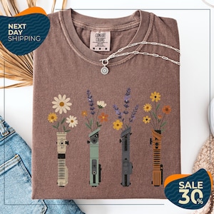 a brown t-shirt with a graphic design of four different colored vases holding flowers, arranged in a row. The vases are depicted in various colors, including green, blue, orange, and brown. The design also includes a small charm or pendant. The t-shirt is displayed on a white background, and there are some jeans and a sweater visible in the background.