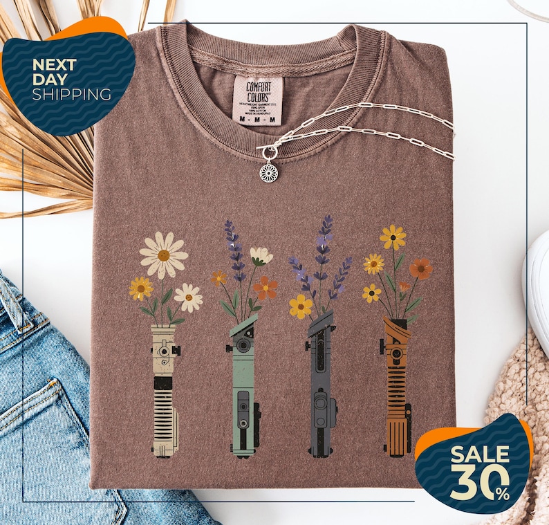 a brown t-shirt with a graphic design of four different colored vases holding flowers, arranged in a row. The vases are depicted in various colors, including green, blue, orange, and brown. The design also includes a small charm or pendant. The t-shirt is displayed on a white background, and there are some jeans and a sweater visible in the background.