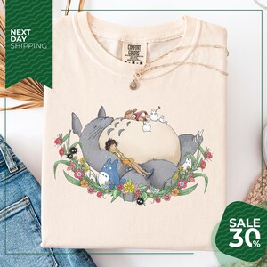 a white t-shirt with a colorful graphic design depicting a cat surrounded by various animals and flowers. The t-shirt is displayed on a white background, and there is a sale sign indicating a 30% discount.