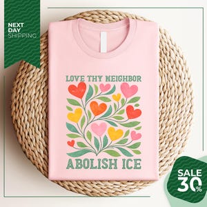 A pink t-shirt with a colorful graphic design of hearts and leaves, along with the text "LOVE thy neighbor ABOLISH ICE" printed on it. The t-shirt is displayed on a woven basket, and there is a green banner with text that reads "NEXT DAY SHIPPING" and "SALE 30%".