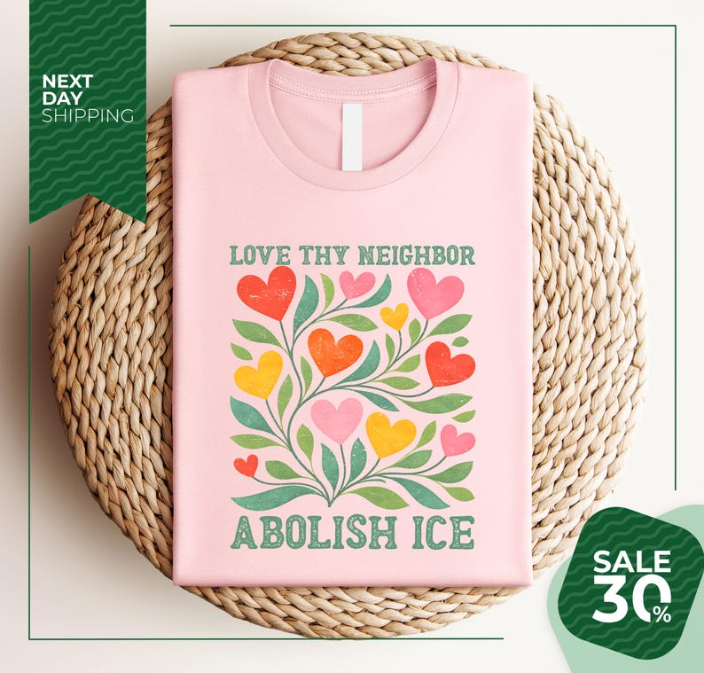 A pink t-shirt with a colorful graphic design of hearts and leaves, along with the text "LOVE thy neighbor ABOLISH ICE" printed on it. The t-shirt is displayed on a woven basket, and there is a green banner with text that reads "NEXT DAY SHIPPING" and "SALE 30%".
