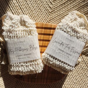 May include: Two white mesh bags containing bars of soap. The soap on the left is labeled "Citrus Hibiscus Bliss" and the soap on the right is labeled "Lavender Fir Essence". Both soaps are labeled "Nourish | Soothe" and have a net weight of 4.5 oz (127g).