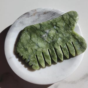 May include: Green jade gua sha tool with a comb on one side. The tool is on a white marble dish.
