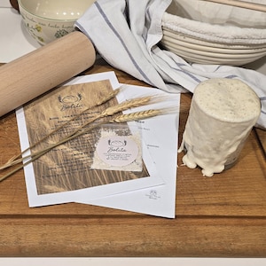 Peut inclure: Un rouleau à pâtisserie en bois, une planche à découper en bois et un pot en verre contenant une substance claire et mousseuse sont disposés sur une surface blanche. Un paquet de farine et des épis de blé sont également présents, ainsi qu'un bol et un linge.