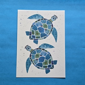 May include: A rectangular white art print featuring two watercolour sea turtles. Each turtle has a shell composed of various shades of blue and green geometric shapes. Small grey speckles are scattered across the white background around the turtles.