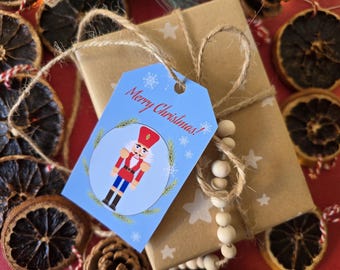 Könnte beinhalten: Ein blaues Geschenkanhänger mit einer Nussknacker-Illustration und dem Text "Frohe Weihnachten!" Der Anhänger ist mit einer Schnur an einer braunen Geschenkbox gebunden, die mit weißen Sternen und einer Holzperlenkette umwickelt ist.