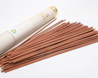 May include: A collection of brown incense sticks next to a beige cylindrical container with gold accents. The container has a label with text and a green and yellow circular logo. The incense sticks are arranged in a pile.