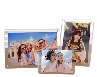 Op de afbeelding: Een verzameling heldere acryl fotolijsten met houten bases. De lijsten tonen verschillende foto's, waaronder een familieportret, twee lachende vrouwen en een portret van een vrouw met een trui en hoed. De lijsten worden bij elkaar gehouden met zilverkleurige hardware.