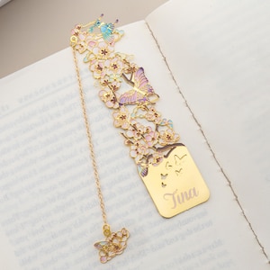 Può includere: Un segnalibro in metallo color oro con un intricato design ritagliato di fiori di ciliegio e farfalle con accenti in smalto rosa pastello, blu e viola. La sezione rettangolare inferiore è incisa con il nome Tina e una piccola catenella.