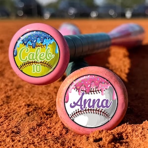 May include: Two baseball bats with pink end caps featuring circular decals. One decal shows a yellow softball with blue icing and sprinkles, labeled Caleb 10. The other shows a white baseball with pink icing and sprinkles, labeled Anna.