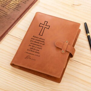 May include: A brown leather journal with a cross and a quote from 1 Thessalonians 5:16-18 engraved on the front. The journal has a strap closure and the name "Bonnie" is written below the quote.
