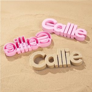 May include: Three sand molds spelling the name "Callie" are displayed on a sandy surface. Two molds are pink plastic, and one is made of sand. The pink molds are in the shape of the name "Callie".