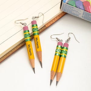 May include: A pair of yellow pencil earrings with green and pink accents. The earrings are personalized with the name "Ms. Harris".