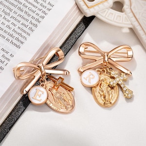 Puede incluir: Dos broches dorados en forma de lazo. Cada broche tiene un respaldo de alfiler y presenta un dije colgante. Uno tiene un círculo blanco con la letra "E", una cruz y un medallón religioso. El otro tiene un círculo blanco con la letra "R", una cruz y un medallón religioso.