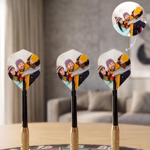 May include: Three dart flights featuring a custom printed photograph of three people wearing winter hats and scarves. The flights are attached to black shafts and gold-colored, textured metal barrels, standing upright on a dartboard.