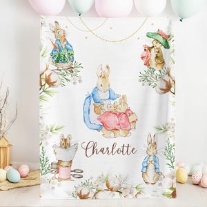 Puede incluir: Una manta de bebé personalizada blanca con un tema de Peter Rabbit. La manta presenta ilustraciones de Peter Rabbit y otros personajes de la historia, incluyendo bolas de algodón, hojas verdes y una cadena dorada. El nombre "Charlotte" está escrito en letras cursivas marrones.