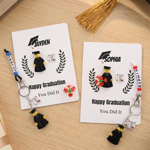 Könnte beinhalten: Zwei Abschlusskarten mit Miniaturfiguren in Abschlusskleidung. Jede Karte trägt einen Namen, "Happy Graduation" und "You Did It". Schlüsselanhänger mit Namen sind angebracht. Einer hat ein blaues Schlüsselband, der andere rot.