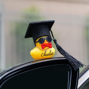 Może przedstawiać: Żółta gumowa kaczka w czapce absolwenta, okularach przeciwsłonecznych i czerwonej muszce. Kaczka ma złoty łańcuszek i imię "Charlotte". Kaczka jest umieszczona na samochodzie, gotowa na ukończenie studiów.