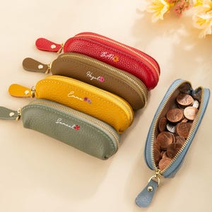 May include: Several personalised leather coin purses in shades of red, brown, yellow, and grey. Each purse features a zip closure and a small wrist strap. The purses are embroidered with names and floral designs.