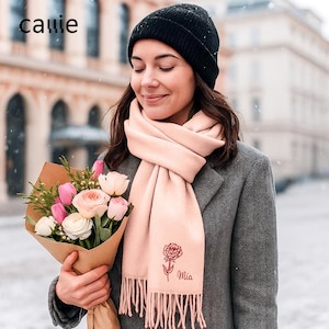 May include: A light pink scarf with a fringed edge and a rose embroidery with the name "Mia". The scarf is draped over a gray coat. A black beanie and a bouquet of flowers are also visible.