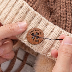 Op de afbeelding: Close-up van een handgemaakte gebreide muts met een bruine leren knoop. De knoop is vastgenaaid met naald en draad. De knoop leest "HANDMADE WITH LOVE BY JENNY". De muts is beige en bruin.
