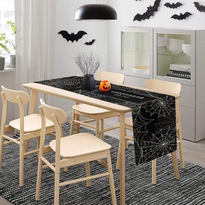 Puede incluir: Un camino de mesa de Halloween en blanco y negro con un diseño de telaraña. El camino de mesa está sobre una mesa de madera con cuatro sillas de madera. También se ve una calabaza tallada y murciélagos decorativos.