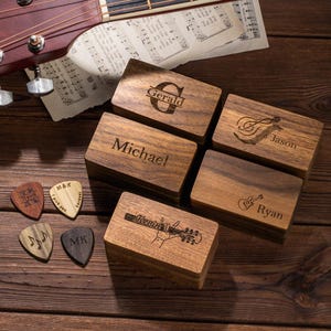 May include: A collection of wooden guitar pick boxes and picks. Each box is engraved with a name, such as "Gerald" and "Michael". The picks have various designs and text, including "I pick you" and musical notes. A guitar headstock is visible.