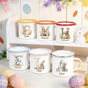 Pode incluir: Canecas de esmalte brancas com bordas coloridas e desenhos de coelhos personalizados. Cada caneca apresenta uma letra, um nome e ilustrações temáticas da Páscoa. As bordas são laranja, vermelha, rosa, azul, amarela e prateada. As canecas estão em uma prateleira branca com decorações de Páscoa.