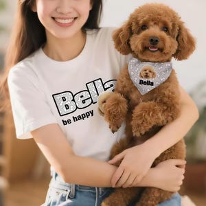 May include: A brown poodle wearing a custom bandana with the name "Bella" and a matching graphic. The dog is being held by a person wearing a white t-shirt with the name "Bella" and the phrase "be happy" printed on it.