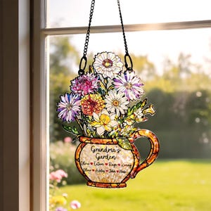 Op de afbeelding: Een gebrandschilderde raamhanger met een boeket kleurrijke bloemen in een decoratieve theekop. De theekop heeft de tekst "Grandma's Garden" en namen. De raamhanger hangt aan een zwarte ketting.