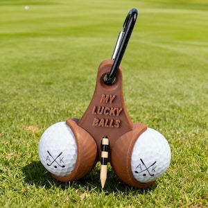 May include: A brown golf ball holder with the text "MY LUCKY BALLS" and a black carabiner clip. Two white golf balls with the letters "A" and "K" and crossed golf clubs are held in place. A golf tee is also included.
