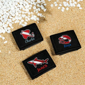 May include: Three black rectangular wallets featuring embroidered red and white marine life designs. One wallet displays a stingray with the name Charles, another a shrimp with the name Freya, and the third a shark with the name James in red text.