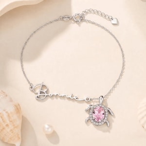 May include: A silver-toned chain bracelet featuring the name Bonnie in cursive script lettering and a turtle-shaped pendant. The turtle charm has a central oval-cut pink gemstone surrounded by a halo of small clear stones and four silver legs.