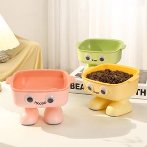 May include: Three colorful pet food bowls with a whimsical design. The bowls are in shades of pink, green, and yellow, each with large, googly-eye details and a pair of stubby legs. One bowl is filled with pet food. The pink bowl has the word "Pudding" printed on it.