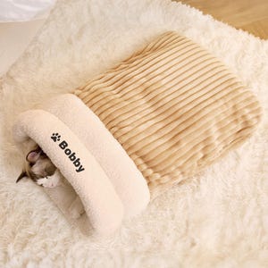 Puede incluir: Una cama para gatos beige con exterior de pana y un interior suave y afelpado. La cama tiene un diseño con capucha, con el nombre "Bobby" impreso en la parte delantera. Un gato está acurrucado dentro de la cama, asomándose por la abertura.